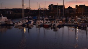 A marina at dusk with boats docked, calm water, and buildings in the background.