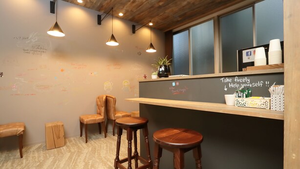 A cozy bar area with wooden stools, a chalkboard with messages, and a shelf with cups and condiments. - bnb+ Tsuruhashi - Hostel (Osaka)
