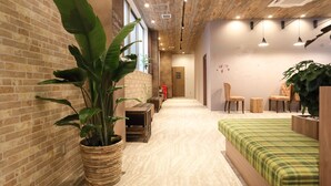 A hotel lobby with a brick wall, large potted plants, a wooden bench, and a bed with a green checkered bedspread. - bnb+ Tsuruhashi - Hostel (Osaka)