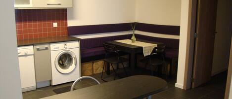 A compact kitchen with a washing machine, a dining table with chairs, and a small table with a vase.