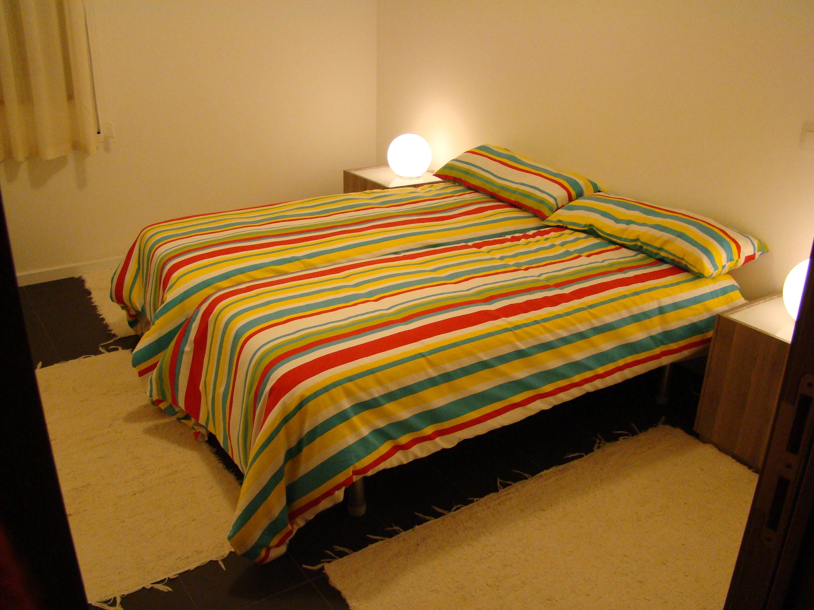 Ein Bett mit gestreifter Bettdecke und Kissen, eine Nachttischlampe und ein Teppich auf dem Boden.