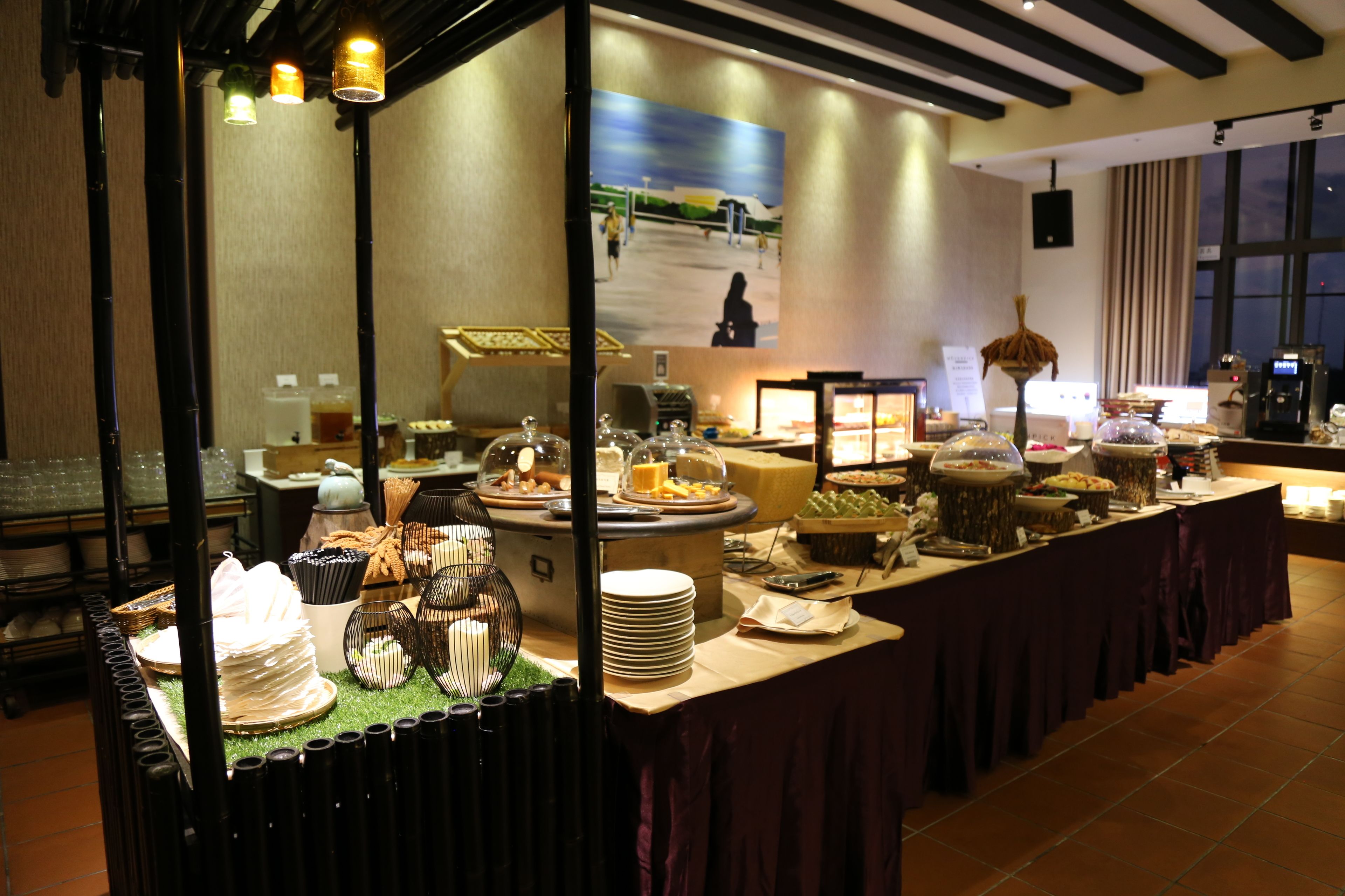 A buffet setup with various food items and serving utensils, a large painting on the wall, and a coffee machine in the background.