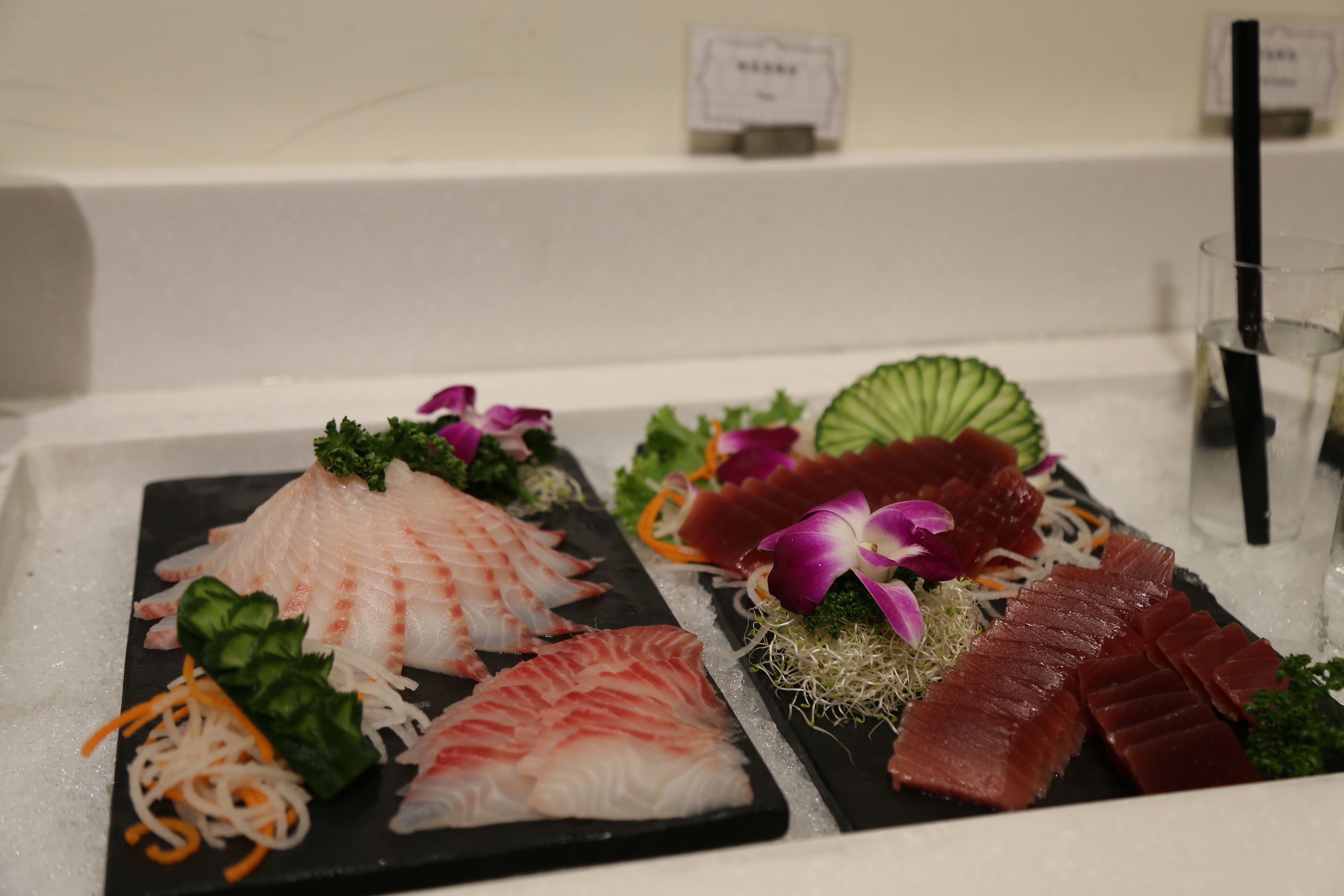 A variety of sashimi on a black slate plate, garnished with green leaves and purple flowers.