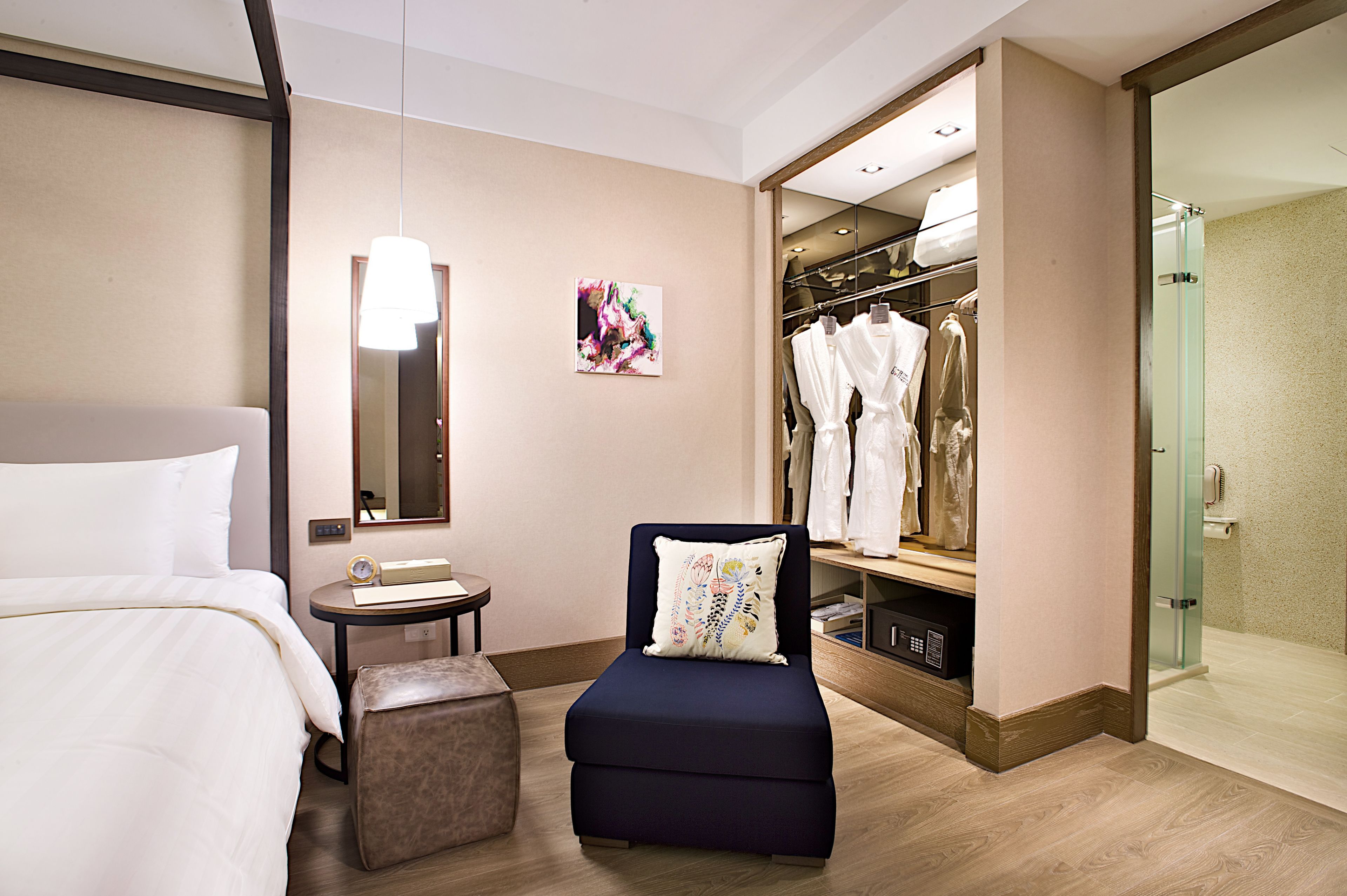 Une chambre d’hôtel comprenant un lit, une petite table avec une horloge, une chaise avec un coussin, une armoire avec des vêtements suspendus et un espace douche.