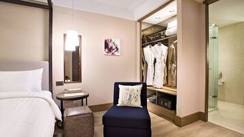 Una habitación de hotel con una cama, una mesita con un reloj, un sillón con una almohada, un armario con ropa colgada y una zona de ducha.