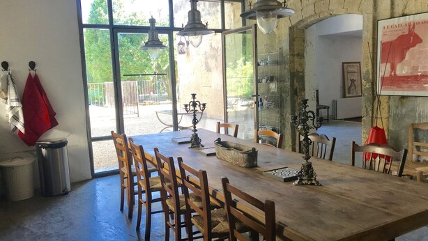 A dining area with a large wooden table, multiple chairs, hanging light fixtures, and a view through large windows to an outdoor patio. - 17th century character house in village center with swimming pool (Saint-Laurent-D'aigouze)