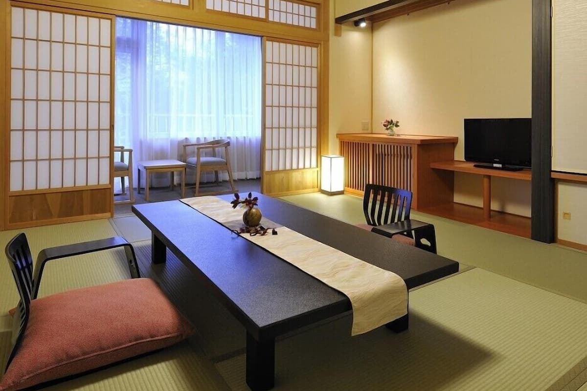 Una habitación tradicional japonesa con suelo de tatami, una mesa baja, sillas, un televisor y puertas correderas.
