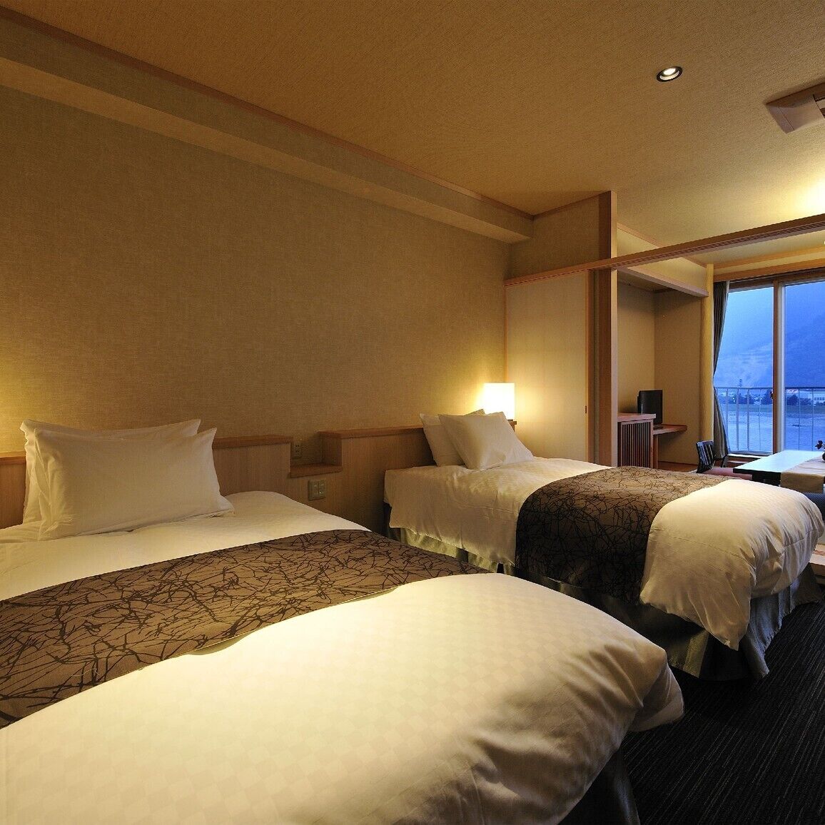 A hotel room with two beds, a desk, and a window with a view.
