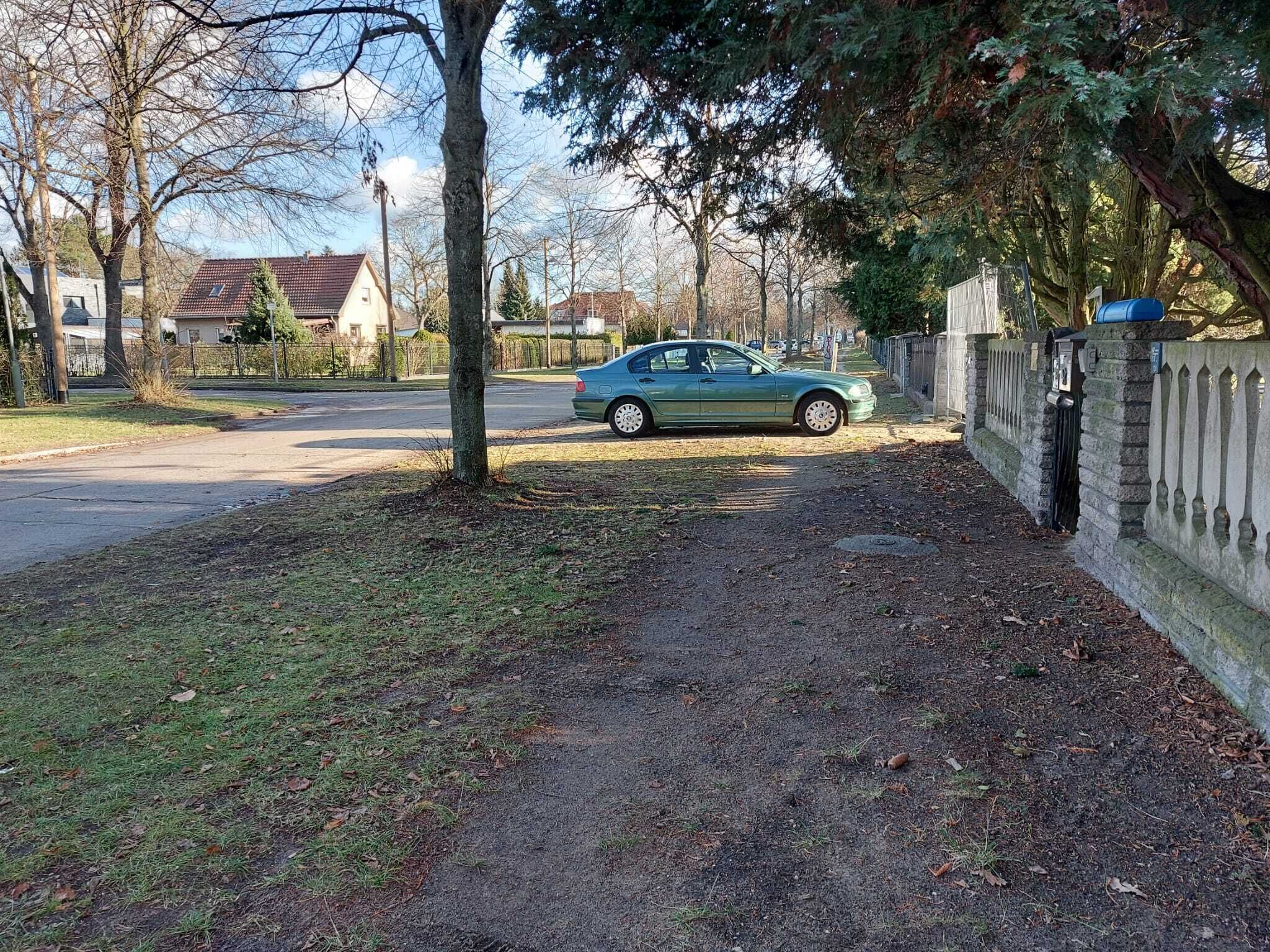 A residential street with a parked car, a tree-lined sidewalk, and houses in the background.