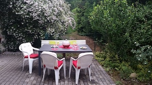 A patio with a table set for four, surrounded by white chairs with red cushions, a bench, and a backdrop of flowering trees. - Beautiful apartment in Berlin-Mahlsdorf (Berlin)