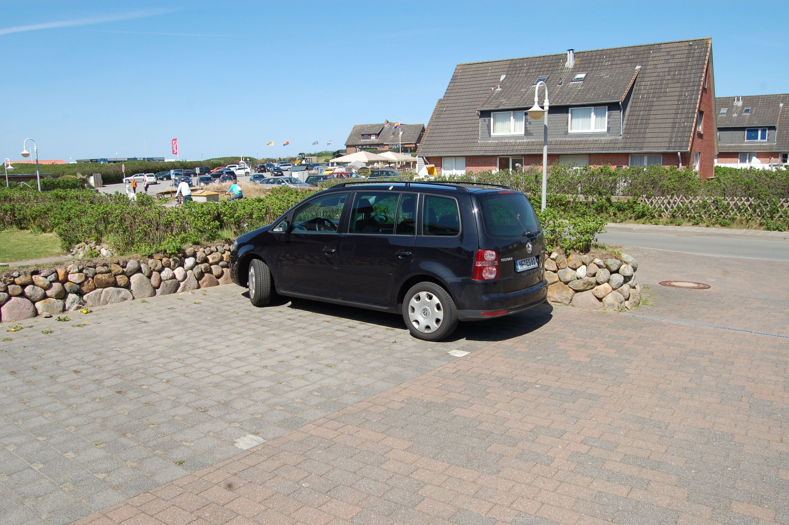 Une voiture noire est garée sur une surface pavée, avec un mur de pierres et un immeuble d’habitation en arrière-plan.