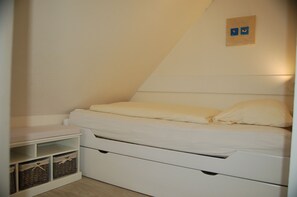 A small, white, single-bed room with a sloped ceiling, a bedside table, and two wicker baskets under the bed.