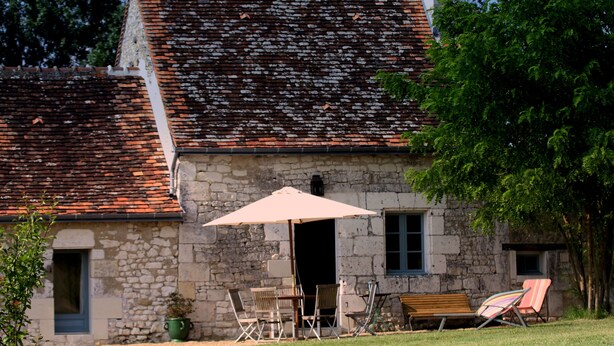 Ein Steinhaus mit einem Ziegeldach, einer Bank und einem Tisch mit Stühlen unter einem Sonnenschirm.