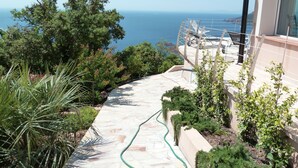 Un camino conduce a una terraza con vistas al mar, rodeada de vegetación y un edificio con balcón.