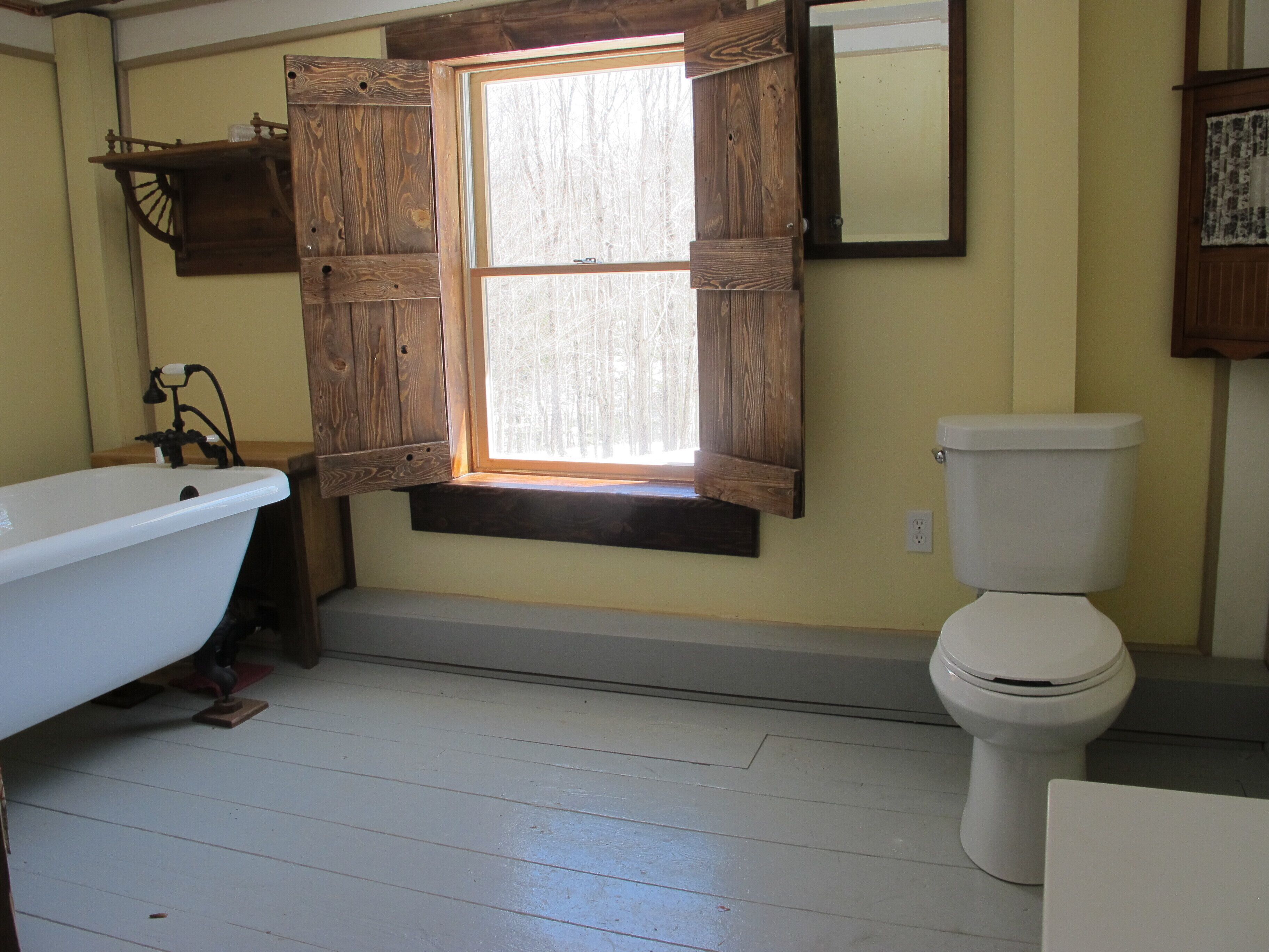 A bathroom with a white bathtub, a toilet, and a window with wooden shutters.
