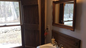 Une salle de bain avec une porte en bois, un lavabo blanc, un miroir et une vue sur un mur de pierre et des arbres à l’extérieur.