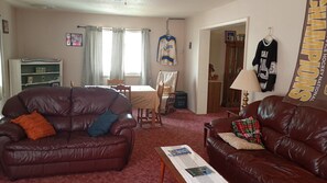 A living room with a brown leather sofa, a dining table with chairs, and a kitchen area visible through an open doorway. - Only 1 dollar cleaning fee at Pictured Rocks Basecamp on the Manistique River! (Germfask)