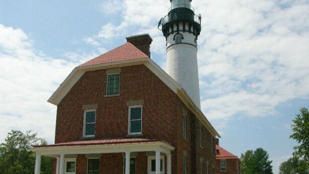 Un bâtiment en briques avec un porche blanc et un phare en arrière-plan.