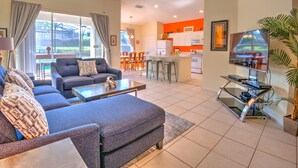 A modern living room with a sofa, ottoman, and coffee table, a dining area with chairs, a kitchen with white cabinets, and a TV on a stand. - Orlando Comfort, Splash and FUN in Windsor Palms Resort - Disney next door (Kissimmee)