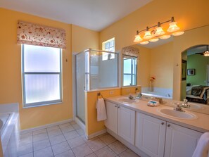 Une salle de bain avec une douche, deux lavabos, un grand miroir et une fenêtre agrémentée de rideaux à motifs floraux.