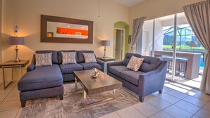 A modern living room with a sectional sofa, a coffee table, and a ceiling fan. There is a view of the outdoors through large windows. - Orlando Comfort, Splash and FUN in Windsor Palms Resort - Disney next door (Kissimmee)