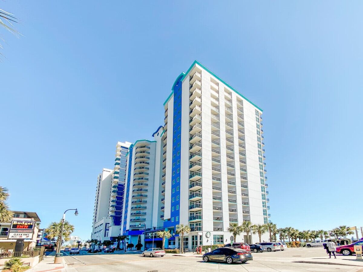 A high-rise hotel with multiple balconies, a clear sky, and a street with cars and palm trees.