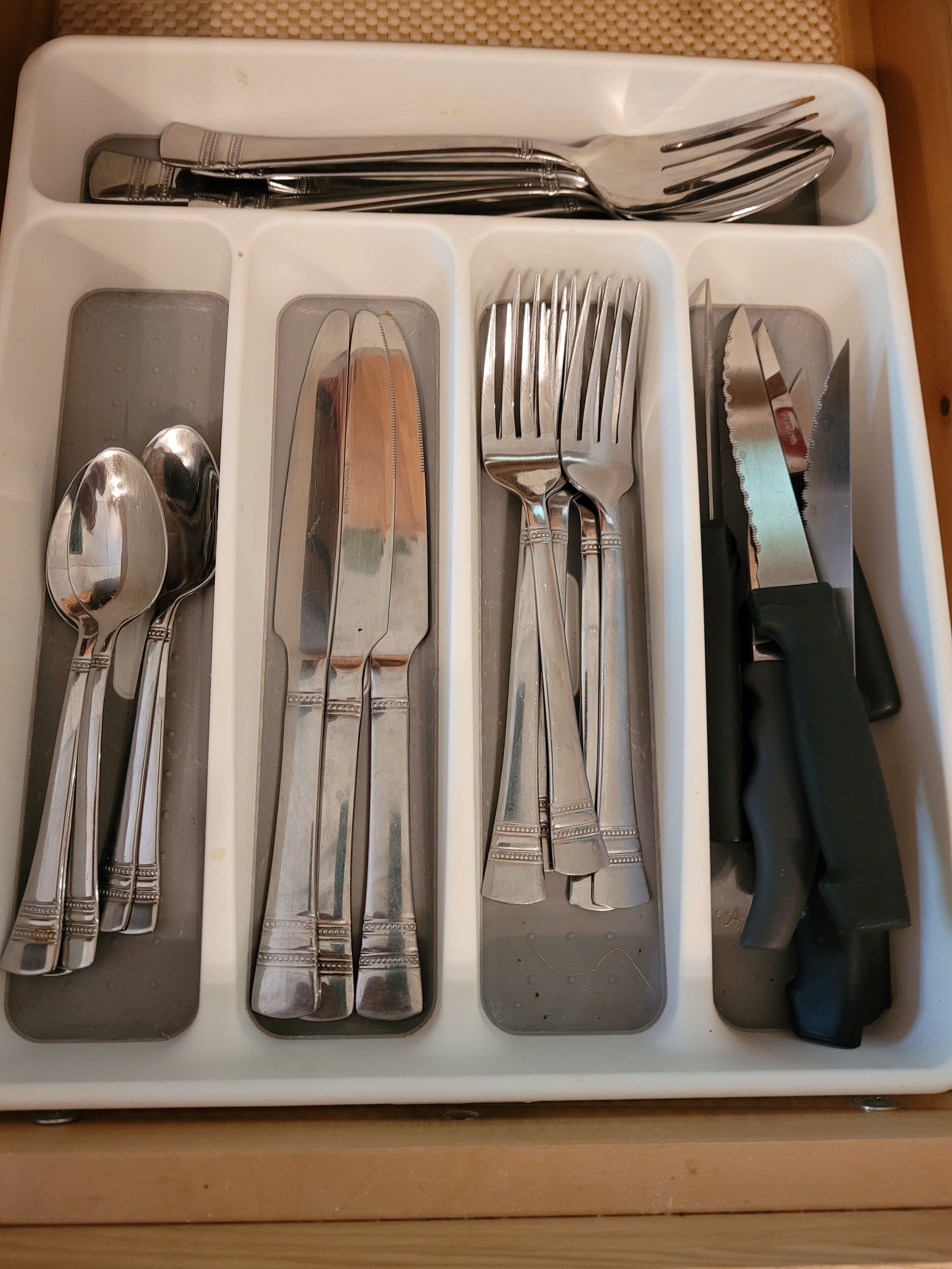 A set of silverware neatly arranged in a white tray with handles, including spoons, a knife, and forks.