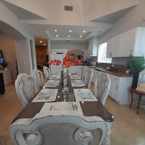 A dining room with a long table set for six, surrounded by chairs. There is a kitchen area with white cabinets and a countertop visible to the right.