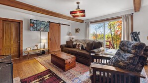 Un salón acogedor con techo de madera, un ventanal con vistas a árboles otoñales, un sofá marrón, un sillón estampado y un aparador de madera.