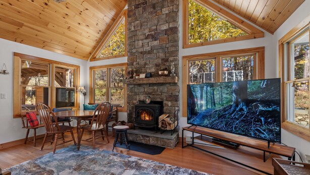 Una sala de estar acogedora con chimenea de piedra, techo de madera, ventanas grandes y un televisor de pantalla plana.