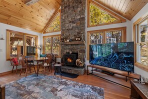 A cozy living room with a stone fireplace, wooden ceiling, large windows, and a flat-screen TV.