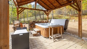 Un área al aire libre con jacuzzi de madera, techo de pérgola, asientos de madera y un hogar para fogata.