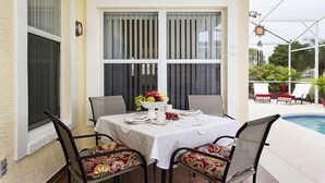 A covered patio with a table set for dining, surrounded by chairs with colorful cushions, and a pool in the background. - Family Disney Villa w/private pool 15 min to Parks (Davenport)