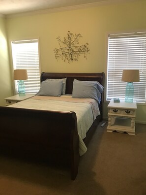 A bedroom with a bed, two bedside tables, a wall decoration, and windows with blinds.