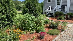 Uma casa com varanda frontal, um jardim com várias plantas e flores, e um gramado.