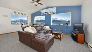 Una sala de estar con un sofá de cuero marrón, una mesa de centro de madera, un televisor sobre un mueble de madera y ventanas grandes con vistas a la montaña.