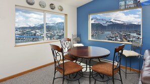 Comedor con mesa redonda y cuatro sillas, ventanas amplias con vistas a un puerto deportivo y montañas, y una pared de acento azul.