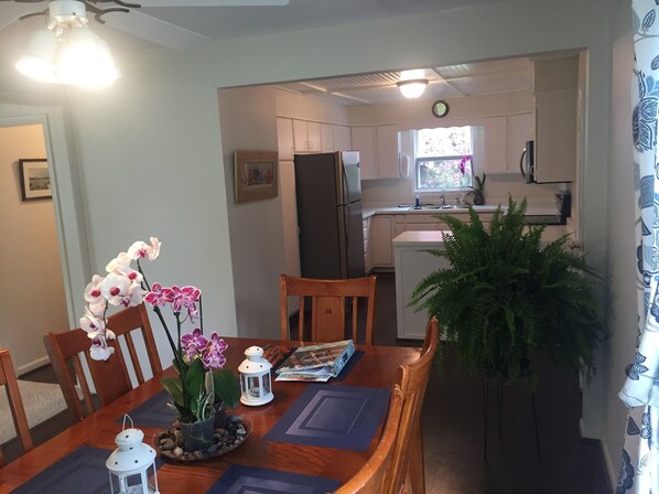 A dining area with a wooden table, chairs, and a potted plant. A refrigerator and kitchen area are visible in the background.