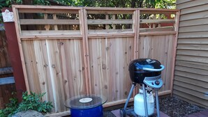 Ein Garten mit Holzpforte, Grill, blauem Fass und Terrassentisch.