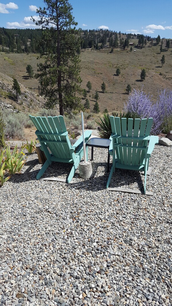 Two green Adirondack chairs and a small table on a gravel surface with a view of a mountainous landscape.