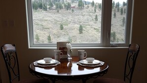 A dining area with a table set for two, a window with a view of a hilly landscape, and a hanging light fixture. - Private Rental Suite: Million Dollar View From Your Deck On 12 Acre Property (Osoyoos)