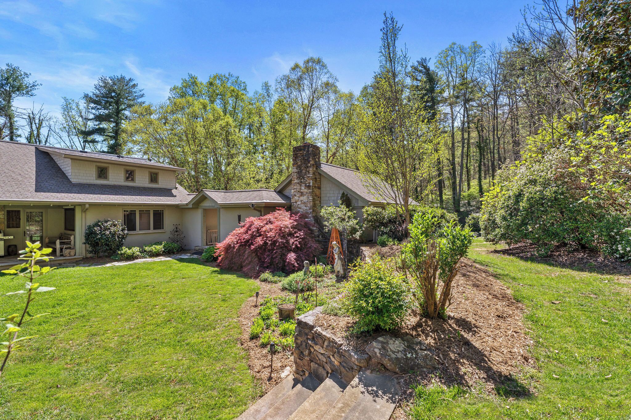 A two-story house with a stone chimney, surrounded by a well-maintained lawn and various shrubs.