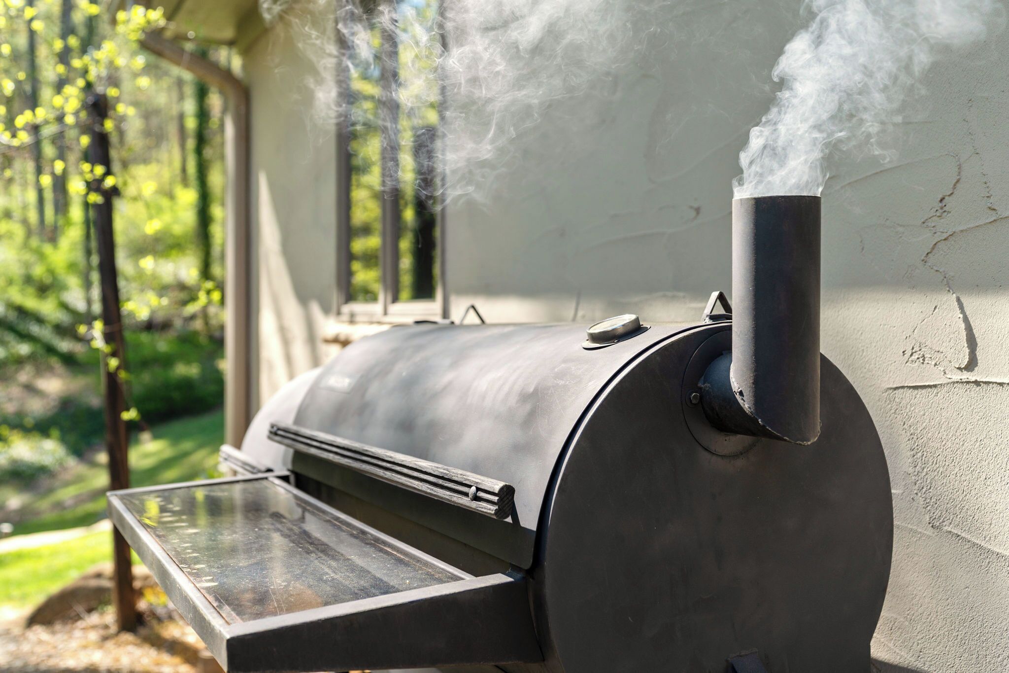 A stainless steel outdoor grill with a chimney, positioned against a wall with a window overlooking a green landscape.
