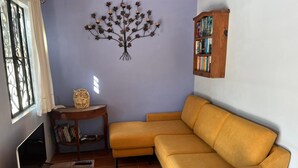 A living room with a sofa, a bookshelf, a decorative tree wall piece, a small table with an owl figurine, and a window with curtains. - Casita Tortuga, spacious 1BR1BA, roof terrace w view, walking dist to beach&town (Sayulita)