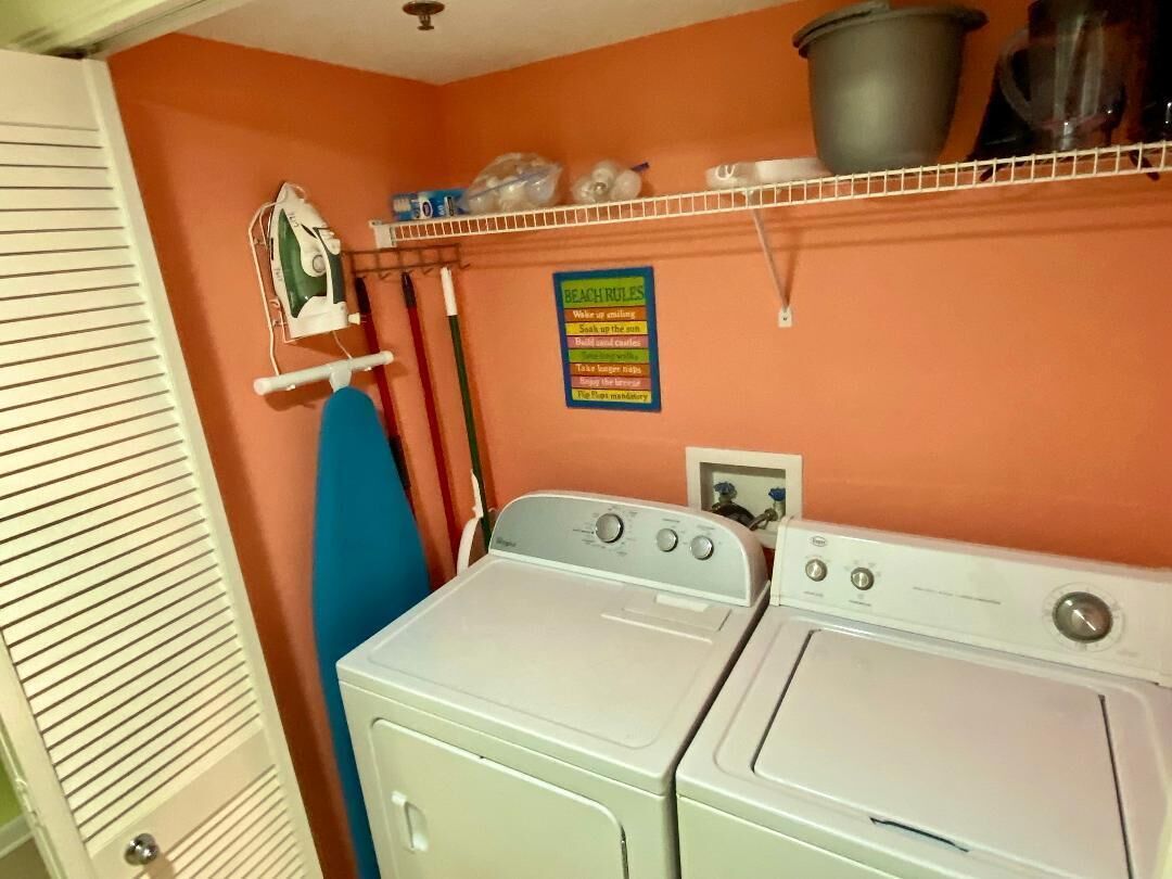 A laundry room with two white front-loading washers and dryers, an ironing board, and a shelf with various items.
