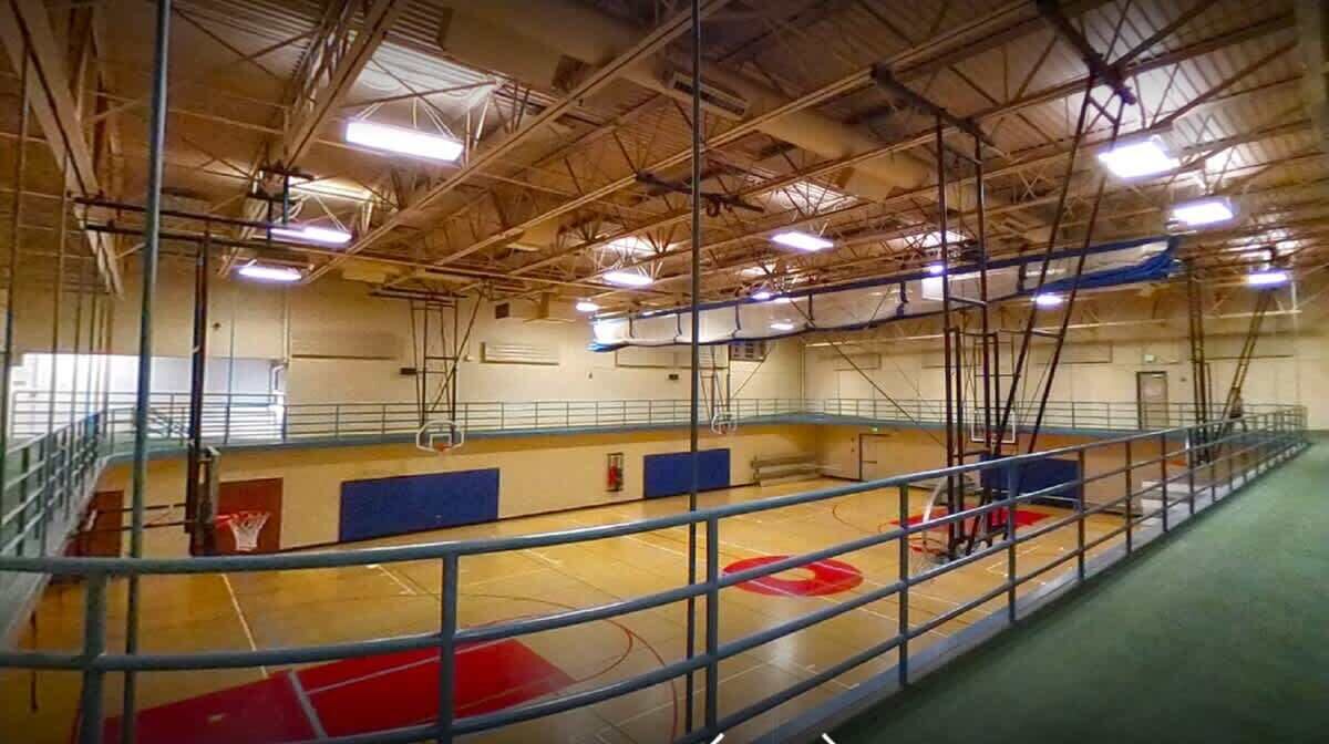 An indoor gymnasium with a basketball court, metal railings, and a high ceiling with visible beams and lighting fixtures.