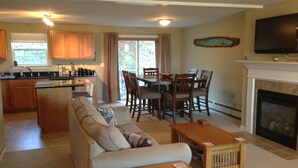 A spacious living room with a sofa, a wooden coffee table, and a fireplace. There is a dining area with a table and chairs, and a kitchen with wooden cabinets and a window. - 3BR/2.5BA Eden Ridge, Great Location for Bethel & Sunday River (Bethel)