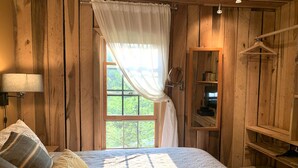 A wooden room with a bed, a window with curtains, a mirror, and a wooden shelf. - Rustic, Artsy & Cozy Getaway Cabin in Laurel Highlands- minutes from Ligonier  (Ligonier)