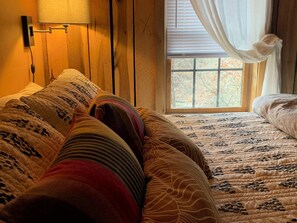 A cozy bedroom with a wooden headboard, a large window with curtains, and a bed with patterned bedding.