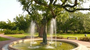 A circular fountain with water jets, surrounded by a brick walkway, set in a park with trees and grass.